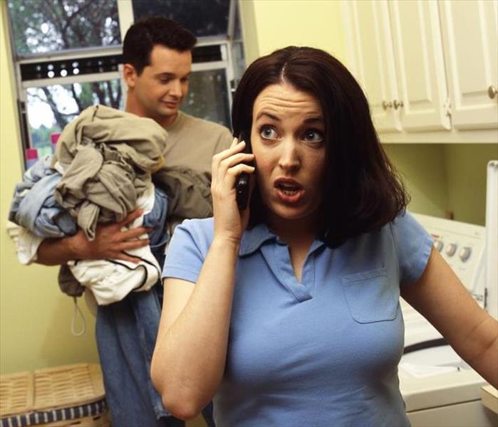 In a laundry room of a house, a woman is on the phone and the man is holding clothes